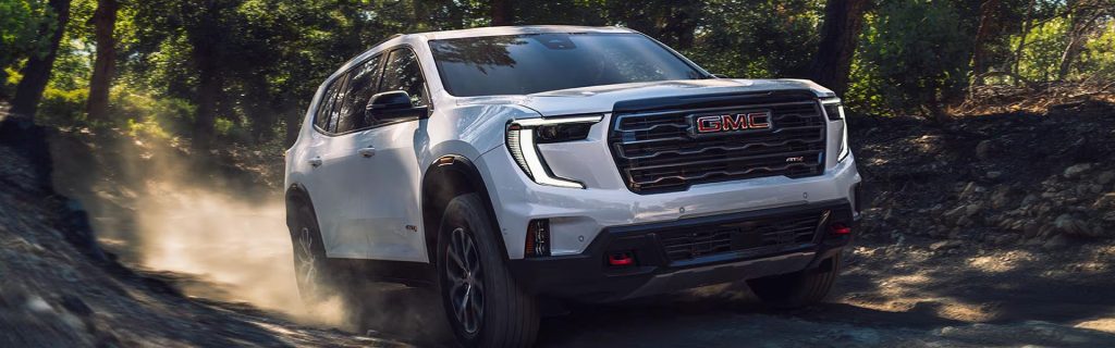 White GMC SUV driving on a dusty forest trail, kicking up dirt as it moves along a rugged, tree-lined path in daylight.
